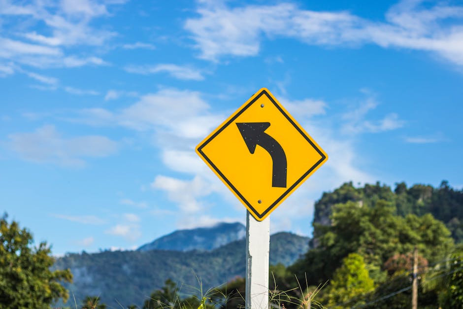 Yellow road sign with a left arrow against a scenic mountain backdrop on a clear day.